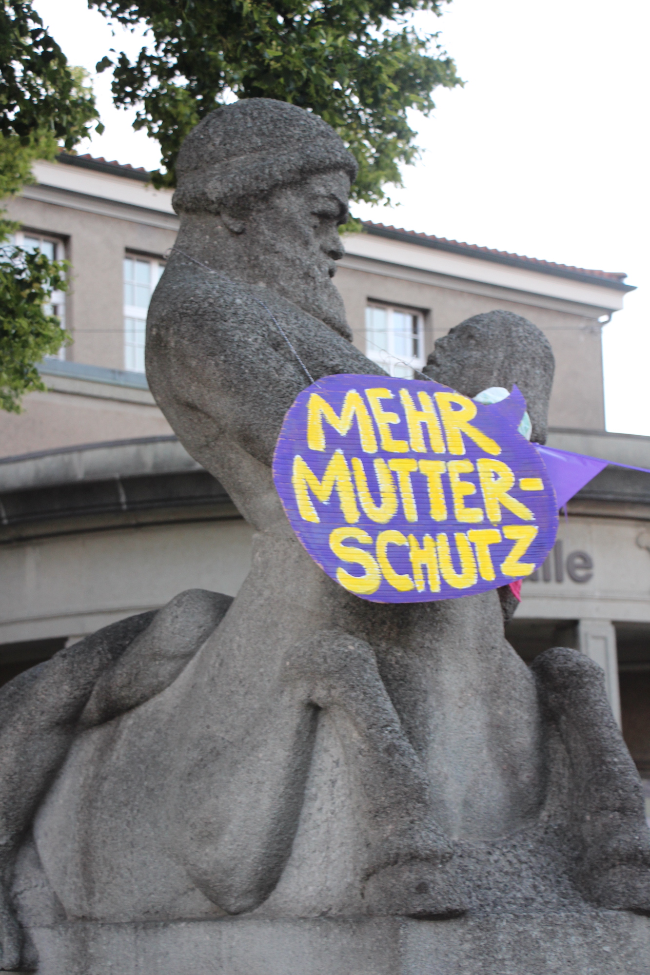 EKdM Kartonschild "Mehr Mutterschutz" an Statue.